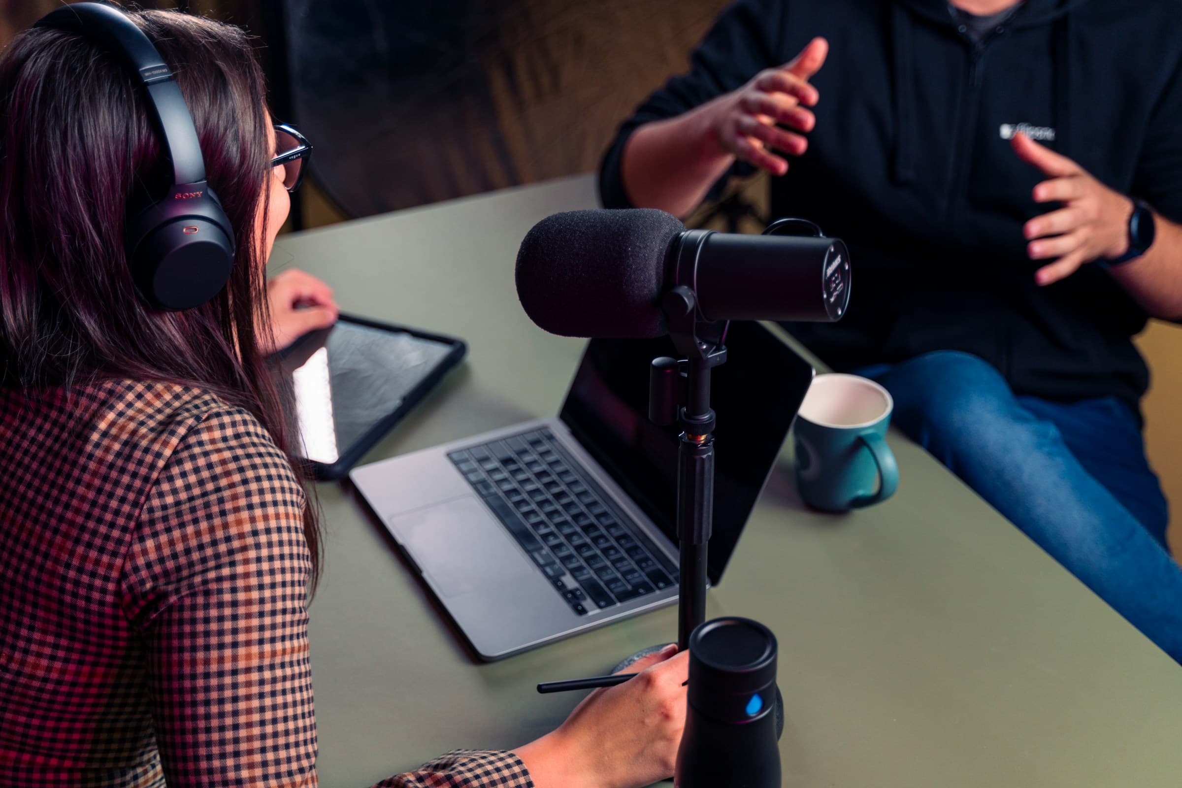 Two people recording a podcast at a table — one wearing headphones and taking notes on a tablet, the other talking and gesturing across a broadcast microphone.