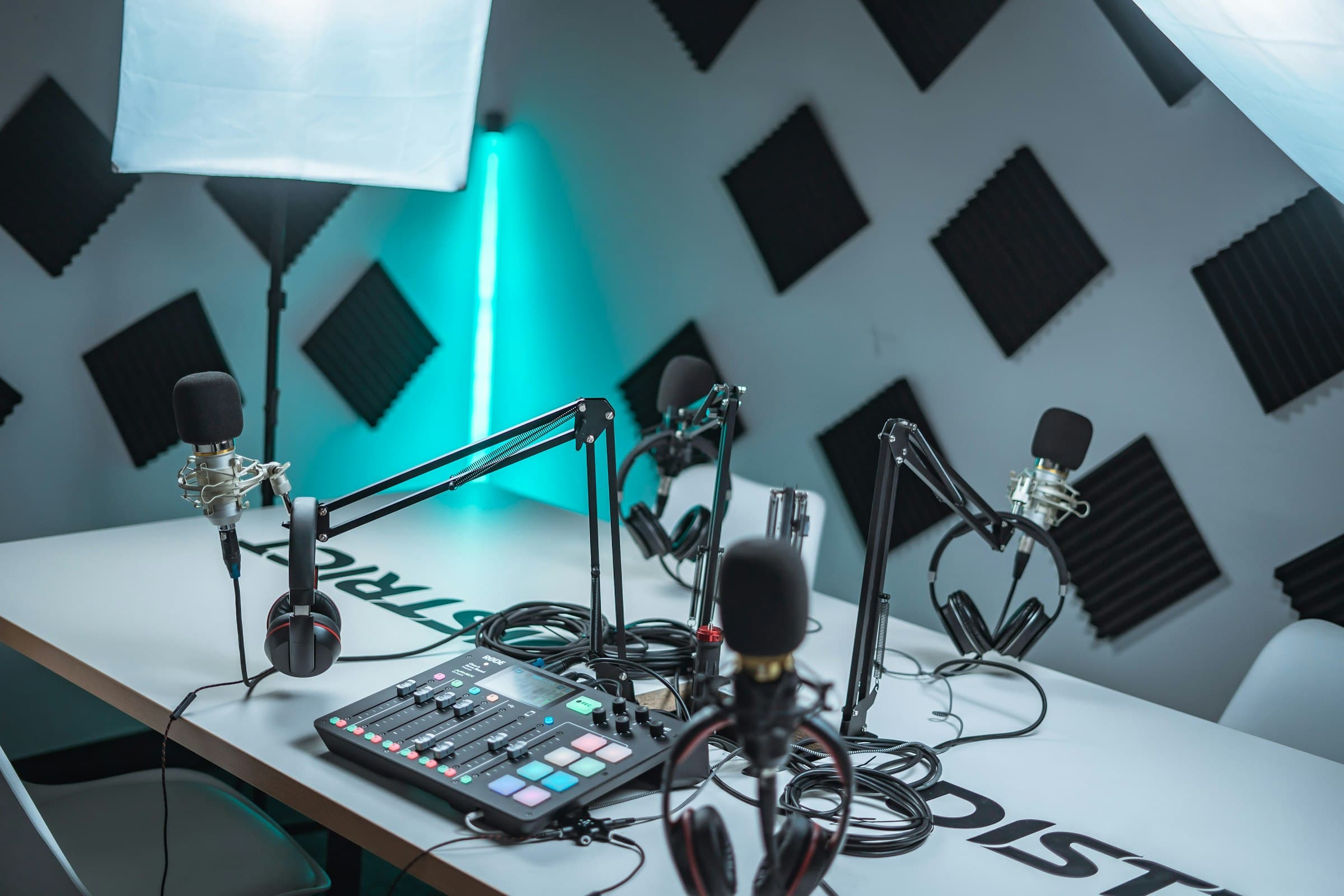 A podcast studio: four microphones on booms, headphones and a mixing console on a white desk, against acoustic foam.