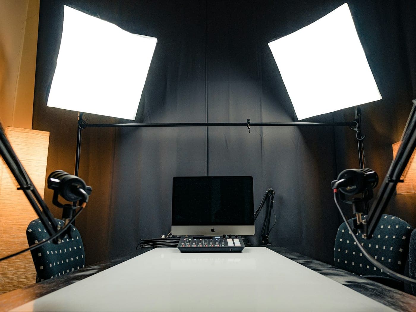 A podcast studio ready to record — two microphones on booms, softbox lights and a mixing desk facing an empty table.