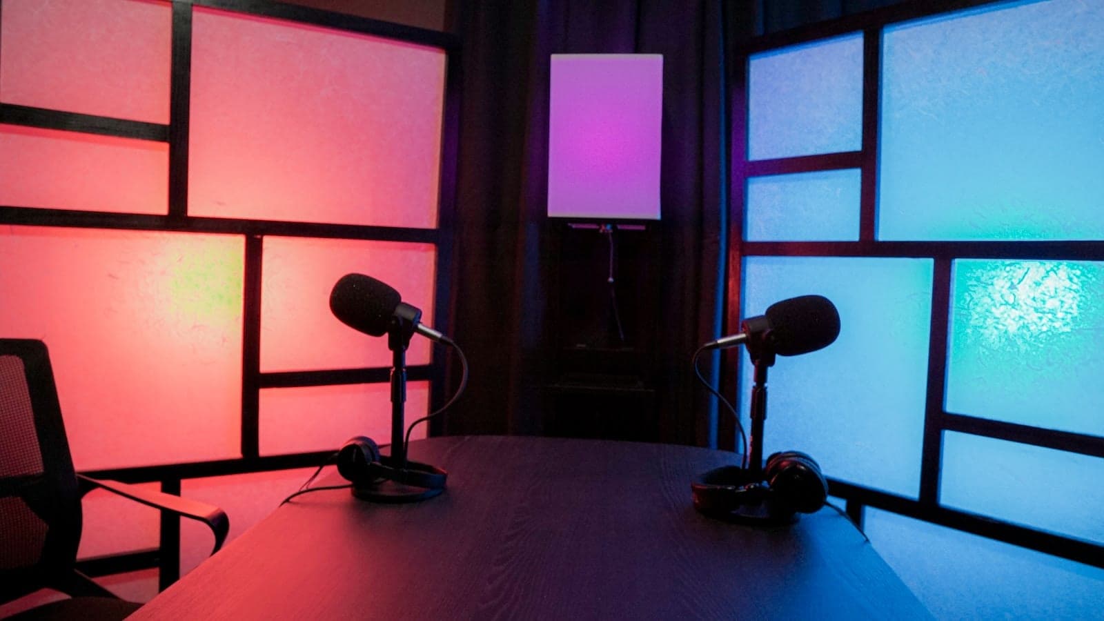 A podcast room lit in red and blue, two broadcast microphones and headphones on a dark table.